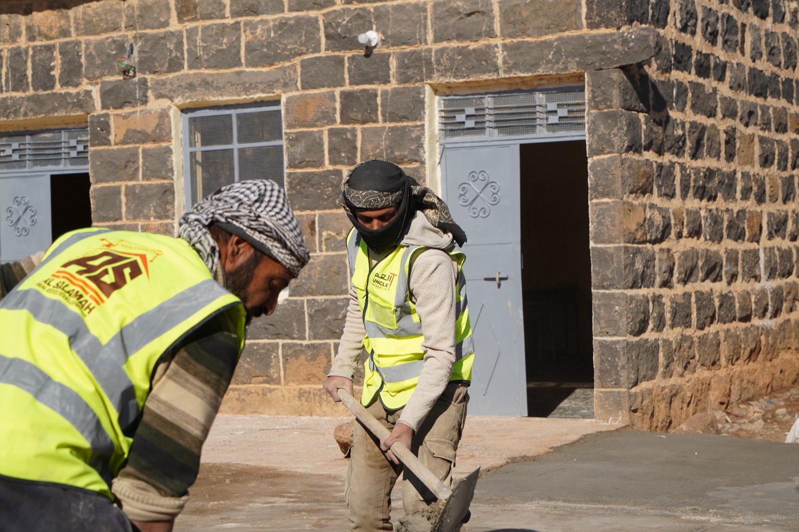 Two Uncle T workers plastering the floor outside a stone school